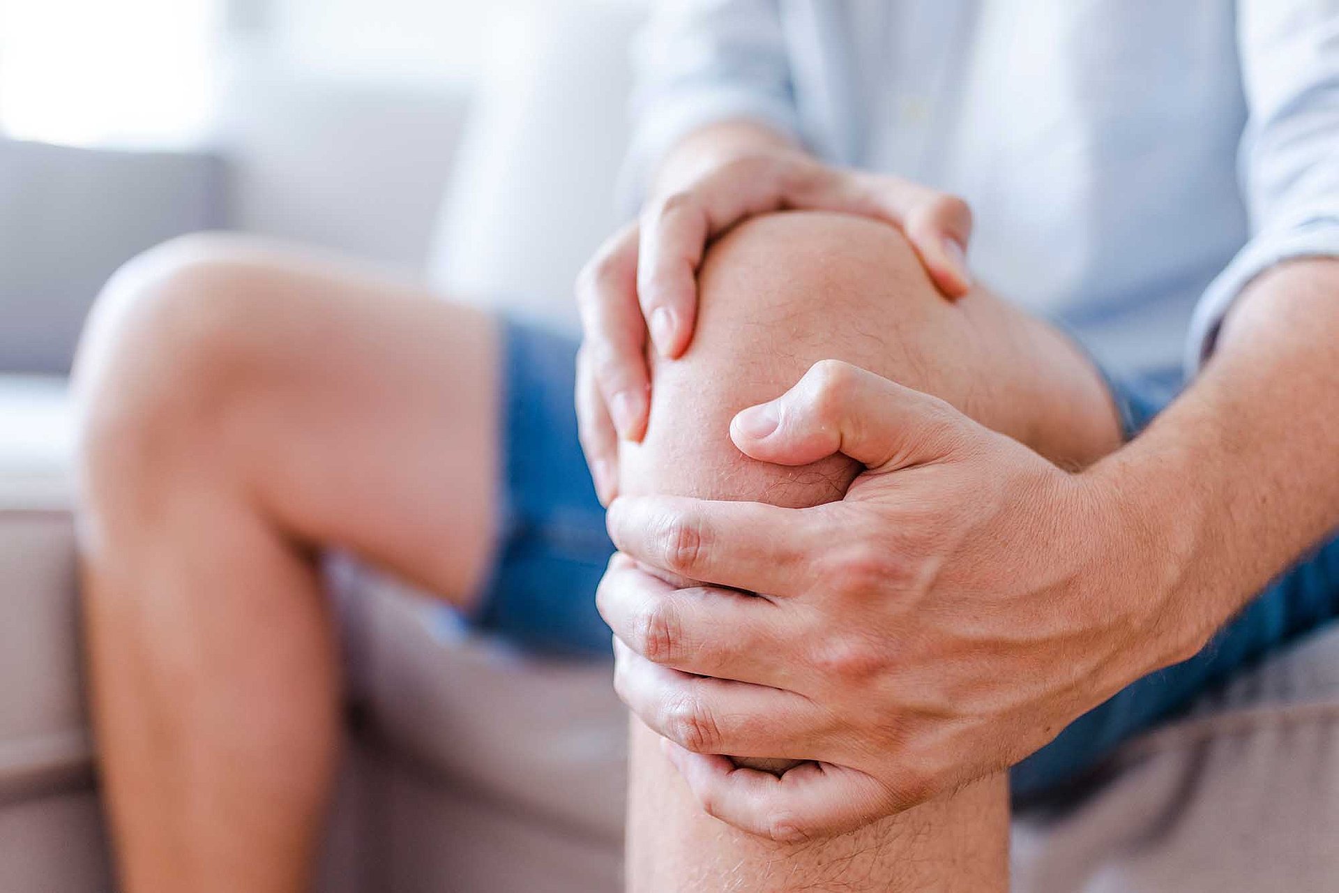 Rohrhof-Apotheke | Arthrose Zu sehen ist ein Bildausschnitt eines Mannes, der auf der Couch sitzend, sein linkes Knie mit beiden Händen festhält. Der Mann hat Schmerzen.